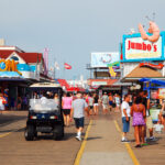 Crowded seaside boardwalk with colorful storefronts and a Jumbo's sign topped by a large shrimp sculpture against a blue sky.