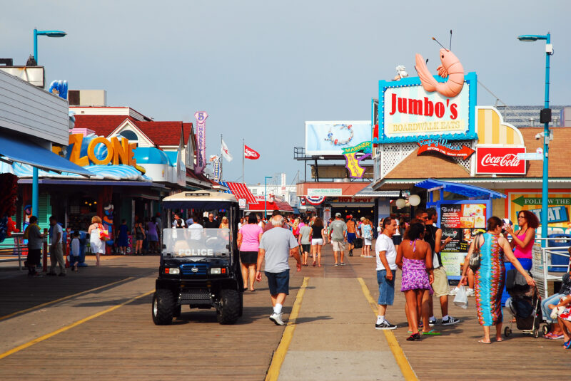Crowded seaside boardwalk with colorful storefronts and a jumbo's sign topped by a large shrimp sculpture against a blue sky.