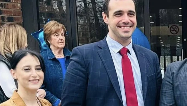 Smiling man in a dark blue pinstripe suit and red tie stands outside with colleagues in front of a glass entrance.