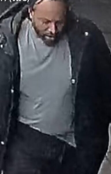 Bearded man in a gray shirt and dark jacket looking down, as if focused or thinking.