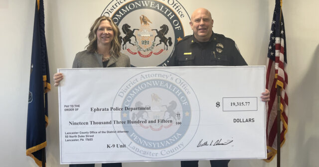 Two officials hold a large ceremonial check for the ephrata police department k-9 unit, $19,315. 77, with a district seal in the background.