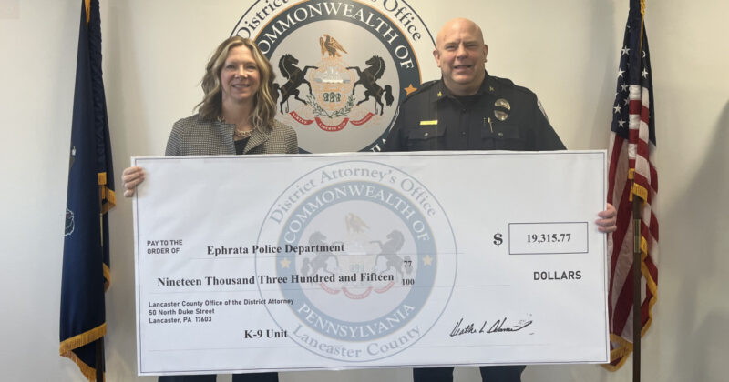 Two officials hold a large ceremonial check for the ephrata police department k-9 unit, $19,315. 77, with a district seal in the background.