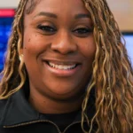Smiling woman with blonde braided hair, wearing a black jacket, in a tech office behind her.