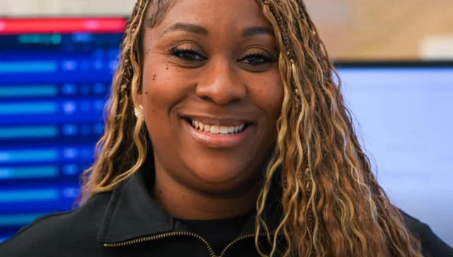 Smiling woman with blonde braided hair, wearing a black jacket, in a tech office behind her.