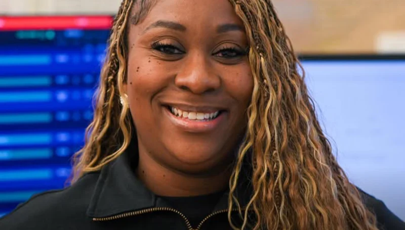 Smiling woman with blonde braided hair, wearing a black jacket, in a tech office behind her.