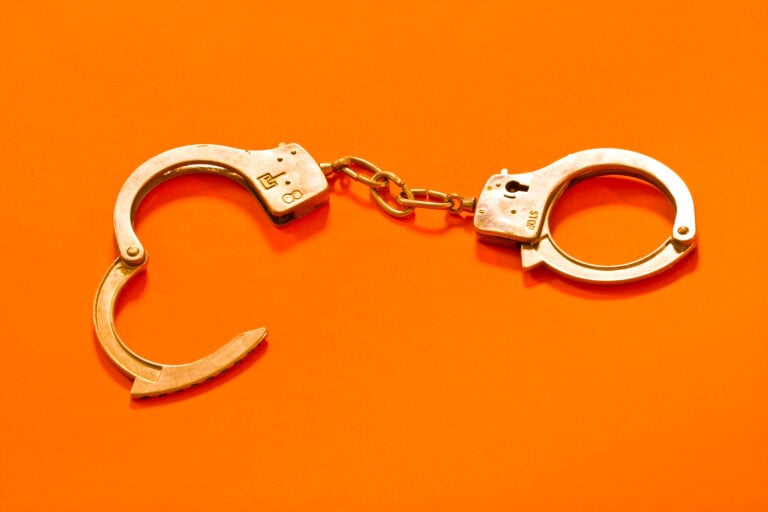 Two metal handcuffs connected by a short chain on a bright orange background; one cuff is open.