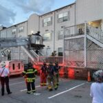 Firefighters on a raised ladder lift work beside a white apartment building at dusk, with onlookers nearby and orange safety cones on the pavement.