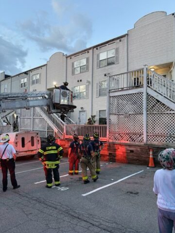 Firefighters on a raised ladder lift work beside a white apartment building at dusk, with onlookers nearby and orange safety cones on the pavement.