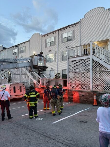 Firefighters on a raised ladder lift work beside a white apartment building at dusk, with onlookers nearby and orange safety cones on the pavement.