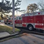 Red fire trucks parked on a suburban street with a utility pole in the foreground during late afternoon; firefighters' equipment visible in the first truck's bay doors.