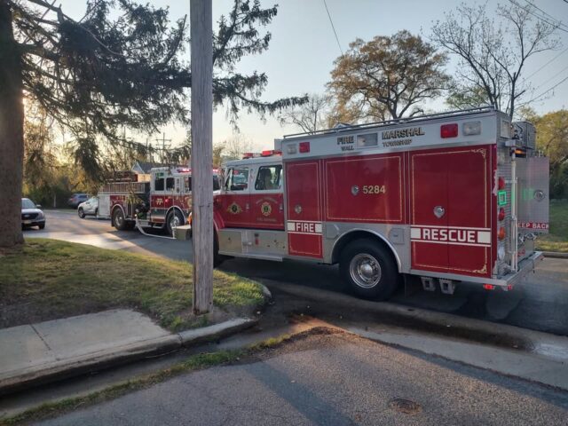 Red fire trucks parked on a suburban street with a utility pole in the foreground during late afternoon; firefighters' equipment visible in the first truck's bay doors.