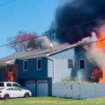 Blue two-story house with white fence on fire; large flames and thick black smoke billow from the right side while a bystander films with a smartphone nearby.