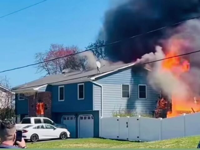 Blue two-story house with white fence on fire; large flames and thick black smoke billow from the right side while a bystander films with a smartphone nearby.