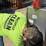 Man in a bright neon shirt leans over and reaches into a tall outdoor metal cabinet on a sidewalk, with boxes and a spray can on top.