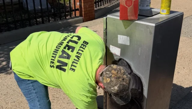 Man in a bright neon shirt leans over and reaches into a tall outdoor metal cabinet on a sidewalk, with boxes and a spray can on top.