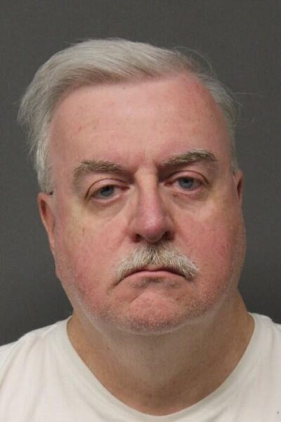 Close-up portrait of an older man with gray hair and mustache, wearing a white shirt, with a neutral expression.