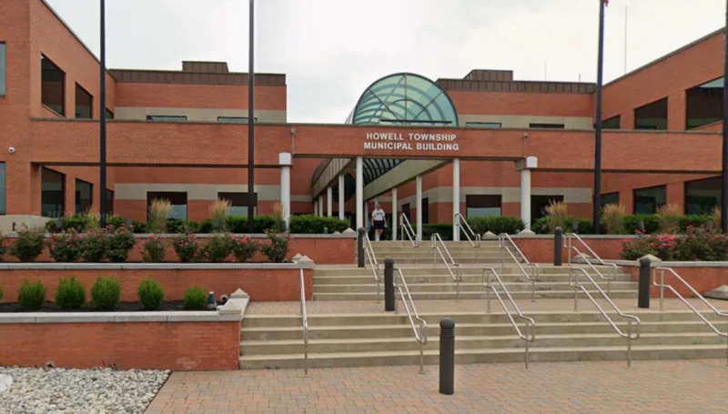 Front of howell township municipal building with brick facade, glass arch, and stepped entrance with metal rails; landscaping on either side.