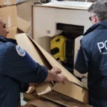 Two police officers wearing HSI jackets inspect a large open crate in a warehouse, revealing a yellow device inside while surrounded by stacked cardboard boxes.