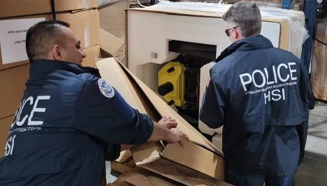 Two police officers wearing hsi jackets inspect a large open crate in a warehouse, revealing a yellow device inside while surrounded by stacked cardboard boxes.