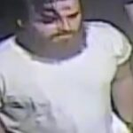 Adult man with dark hair and mustache wearing a white short-sleeve shirt, standing in a dim indoor setting.