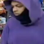 Person in a purple hoodie with the hood up and face partially covered by a black scarf, holding a small blue object in a store aisle; police/crime alert logos partially visible at the top corner
