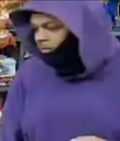Person in a purple hoodie with the hood up and face partially covered by a black scarf, holding a small blue object in a store aisle; police/crime alert logos partially visible at the top corner