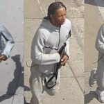 Suspect in a light gray hooded tracksuit walks on a sidewalk, holding a long black firearm.