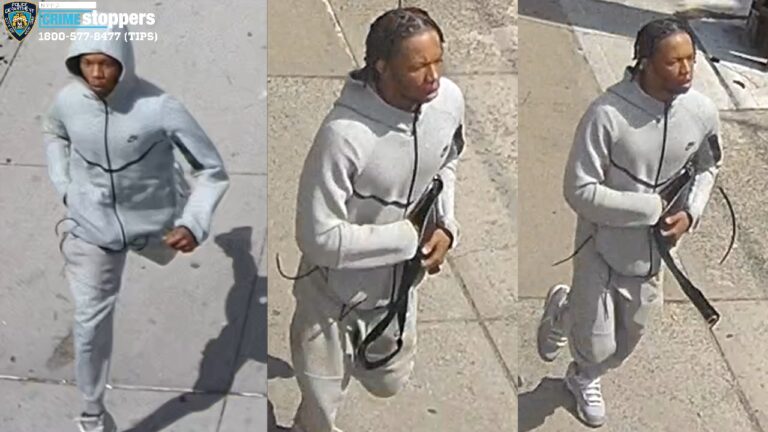 Suspect in a light gray hooded tracksuit walks on a sidewalk, holding a long black firearm.