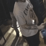 Bearded man wearing a red knit cap and gray T-shirt standing at a dim bar, looking down and holding a drink.