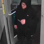 Person in a black hoodie and glasses stepping through a doorway at a transit station with a barrier arm nearby.