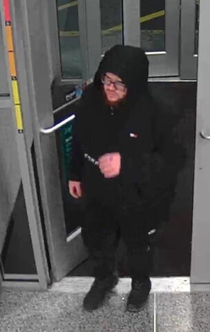 Person in a black hoodie and glasses stepping through a doorway at a transit station with a barrier arm nearby.