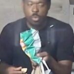Man in a dark shirt sits indoors, holding a green patterned bag and a small snack in his hands, with shopping bags nearby.