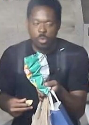 Man in a dark shirt sits indoors, holding a green patterned bag and a small snack in his hands, with shopping bags nearby.