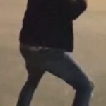 Man in a dark jacket and jeans mid-motion in a parking lot at night, arms raised as if dancing, with cars in the background.