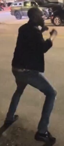 Man in a dark jacket and jeans mid-motion in a parking lot at night, arms raised as if dancing, with cars in the background.