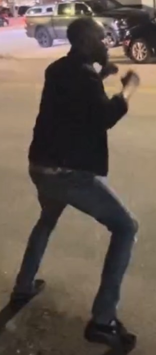 Man in a dark jacket and jeans mid-motion in a parking lot at night, arms raised as if dancing, with cars in the background.