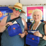 Two attendees at a Maryland Lottery booth pose for a photo, each holding a blue prize bag and smiling at the camera with drinks in hand.