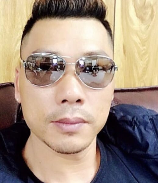 Man wearing reflective aviator sunglasses looking at camera, with short dark hair and a black shirt in a casual setting.