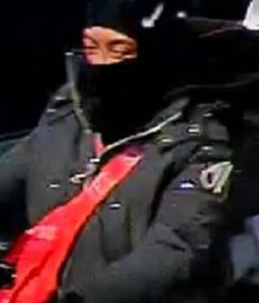 Person wearing a black coat with a bright red scarf, face partially covered by a black veil/mask, with only the eyes visible.