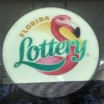 Circular Florida Lottery sign featuring a pink flamingo and teal 'Lottery' text, hanging indoors.