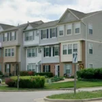 Row of three-story townhomes with beige/gray siding, brick accents, and landscaped lawns in a residential area.