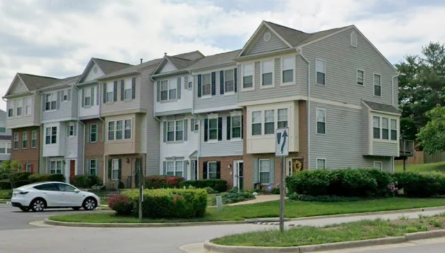 Row of three-story townhomes with beige/gray siding, brick accents, and landscaped lawns in a residential area.
