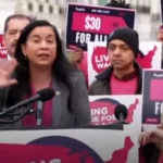 Woman at a protest podium speaks into a microphone as supporters around her hold pink '$30 for all' signs behind.