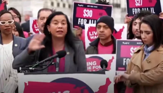 Woman at a protest podium speaks into a microphone as supporters around her hold pink '$30 for all' signs behind.
