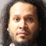 Close-up portrait of a man with long, curly dark hair and a trimmed beard, looking straight ahead against a plain gray background.