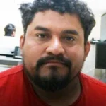Close-up portrait of a man with dark hair and a beard wearing a red shirt, looking at the camera in an office setting.