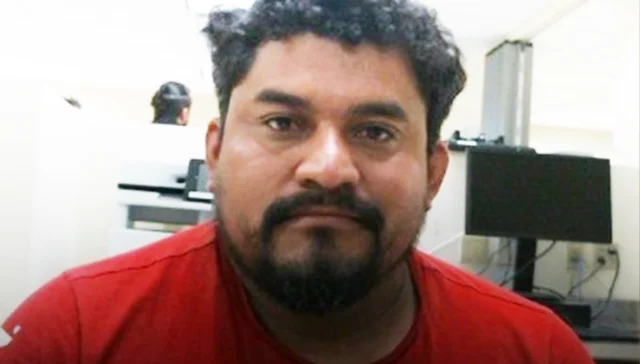 Close-up portrait of a man with dark hair and a beard wearing a red shirt, looking at the camera in an office setting.