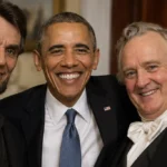 Three men in tuxedos posing together at a formal event, smiling for the camera, with the center man wearing a suit and blue tie and an American flag pin on his lapel.
