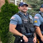 Two uniformed police officers standing outdoors by a brick building, wearing blue shirts, tactical vests, and caps with patches on sleeves.