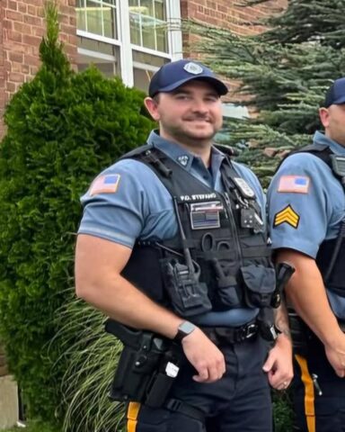 Two uniformed police officers standing outdoors by a brick building, wearing blue shirts, tactical vests, and caps with patches on sleeves.